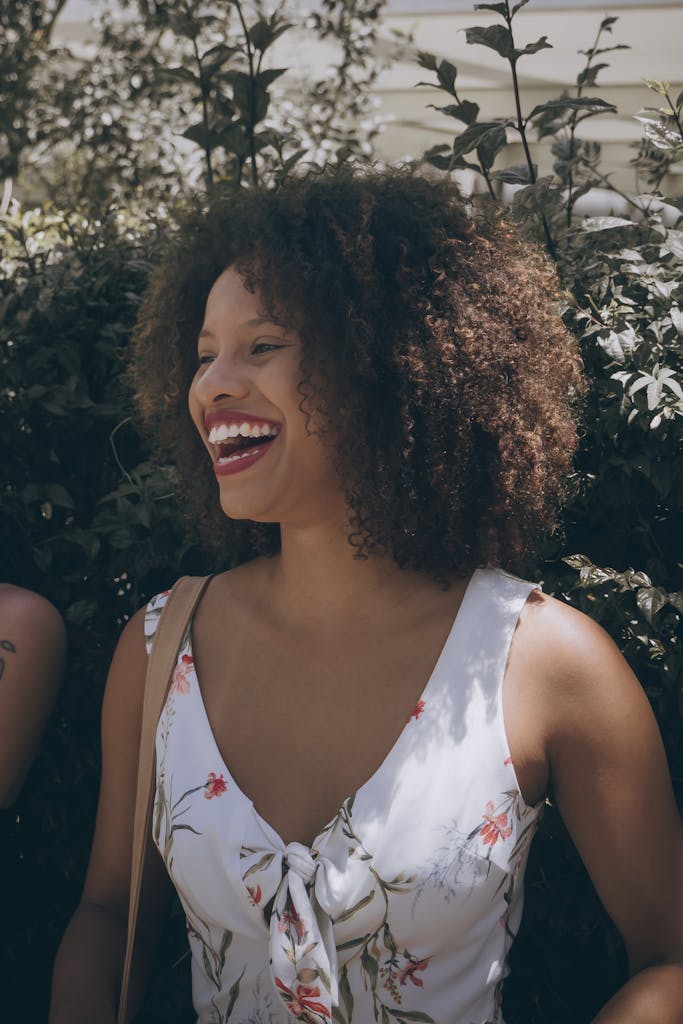 A cheerful woman with curly hair laughing outdoors on a sunny day, in a floral dress.
